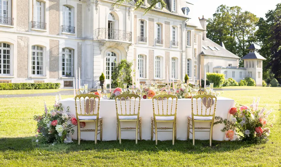 A long banquet table set on the lawn in front of a chateau facade, with floral runners and gilded chairs