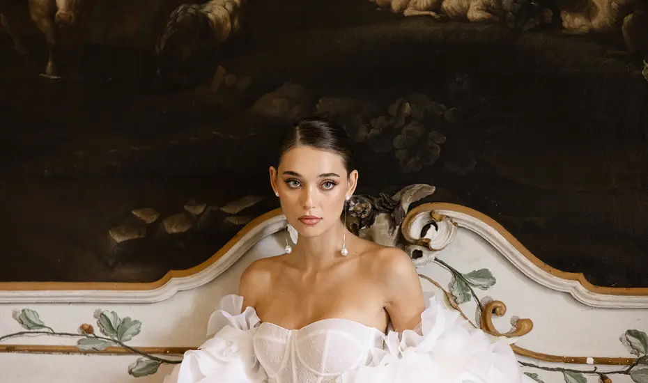 A bride in a white off-the-shoulder dress resting against an antique painted wood panel
