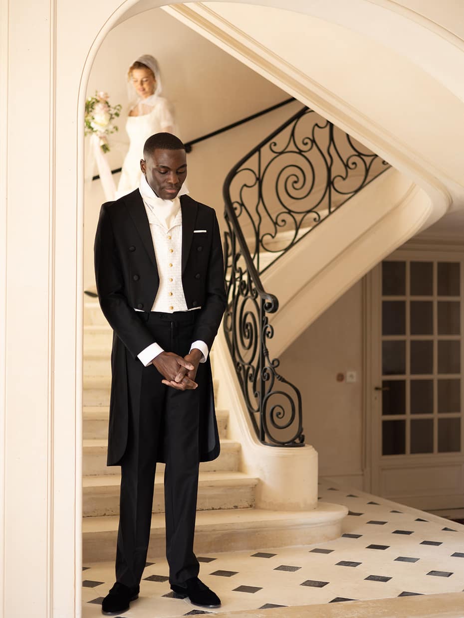 A groom in tails waiting at the foot of a stone staircase with a wrought iron banister, the bride coming down the steps behind him