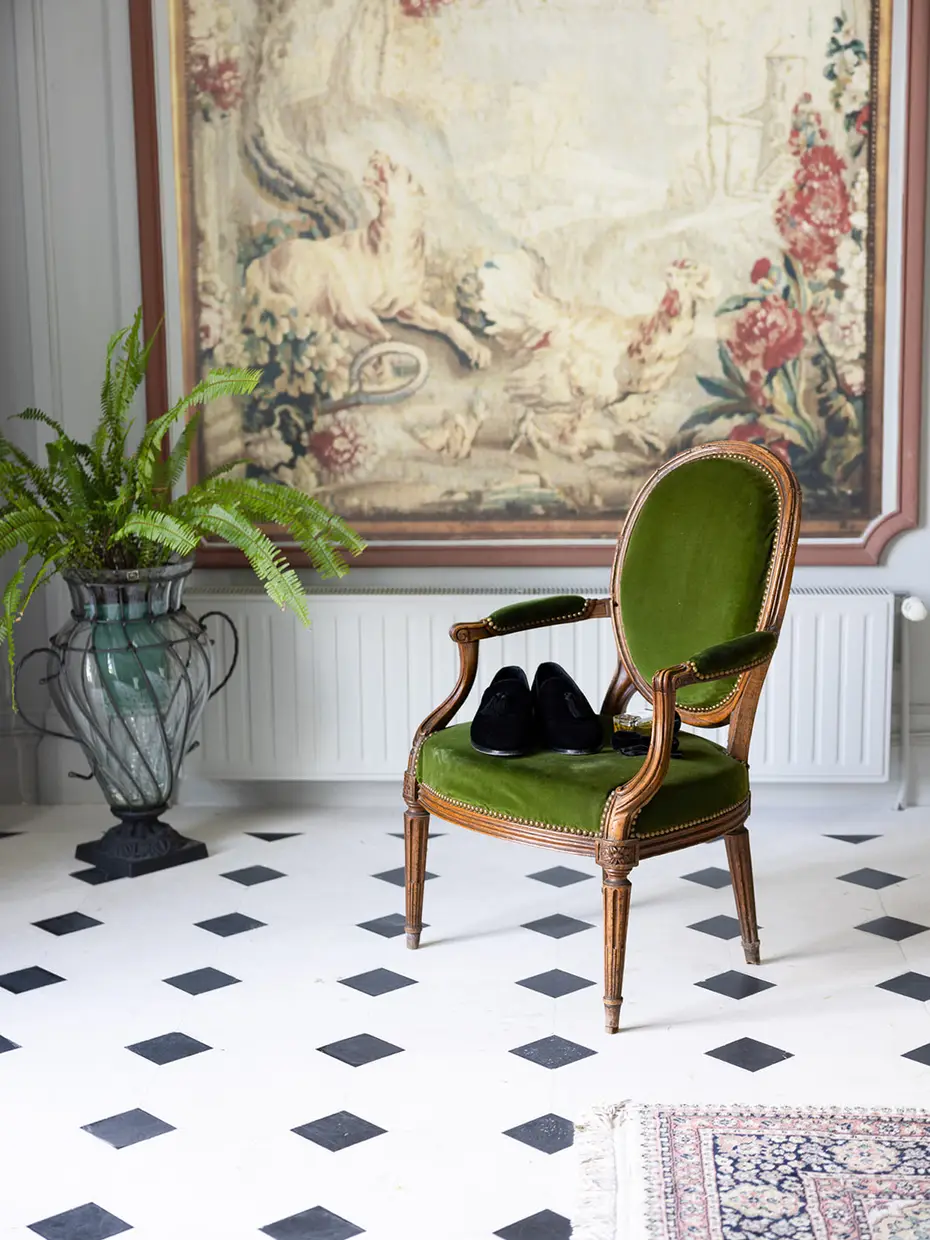 A pair of heels resting on an antique armchair in a room with a chequerboard floor, in front of a wall tapestry