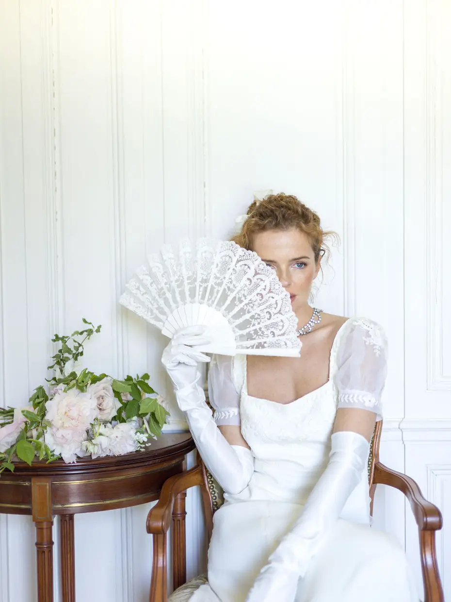 A bride in long gloves holding a lace fan near her face, seated beside pale panelling