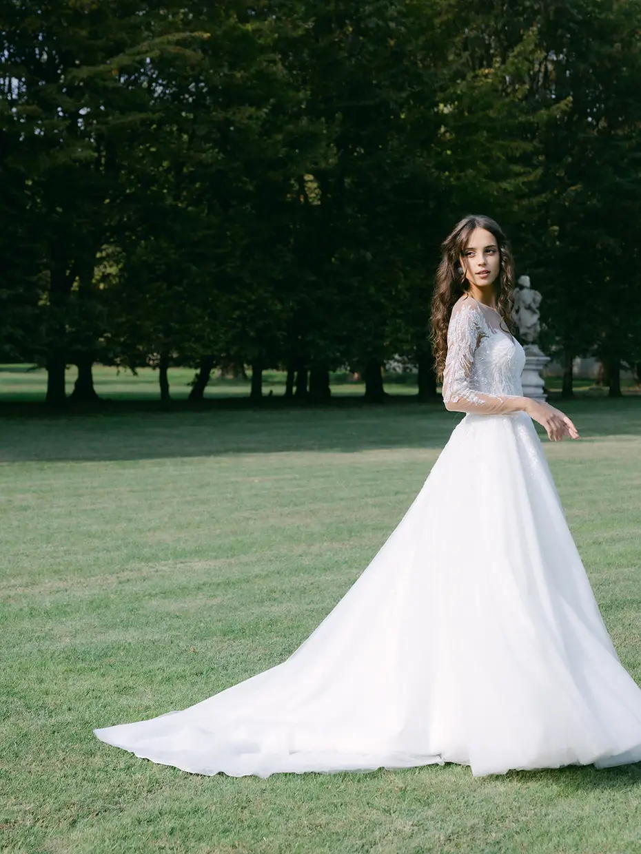 A bride walking alone across a lawn lined with tall trees, in a long-sleeved gown with a full skirt