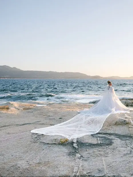 A bride standing on the rocks at the water's edge, her long lace train spread across the stone
