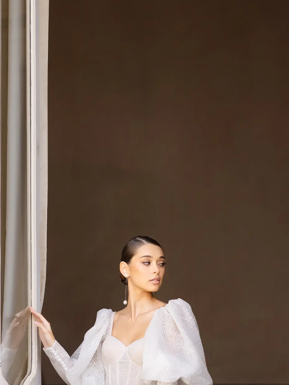 Studio portrait of a bride in a puff-sleeved gown, drawing back a pale curtain against a warm brown backdrop