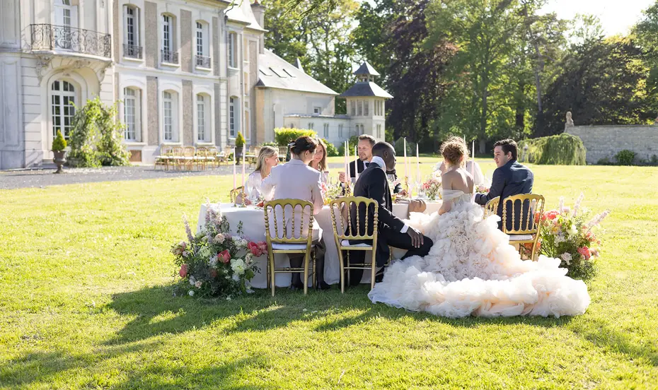 The couple and their guests seated at a round table on the lawn in front of a chateau, the train of the dress spread over the grass