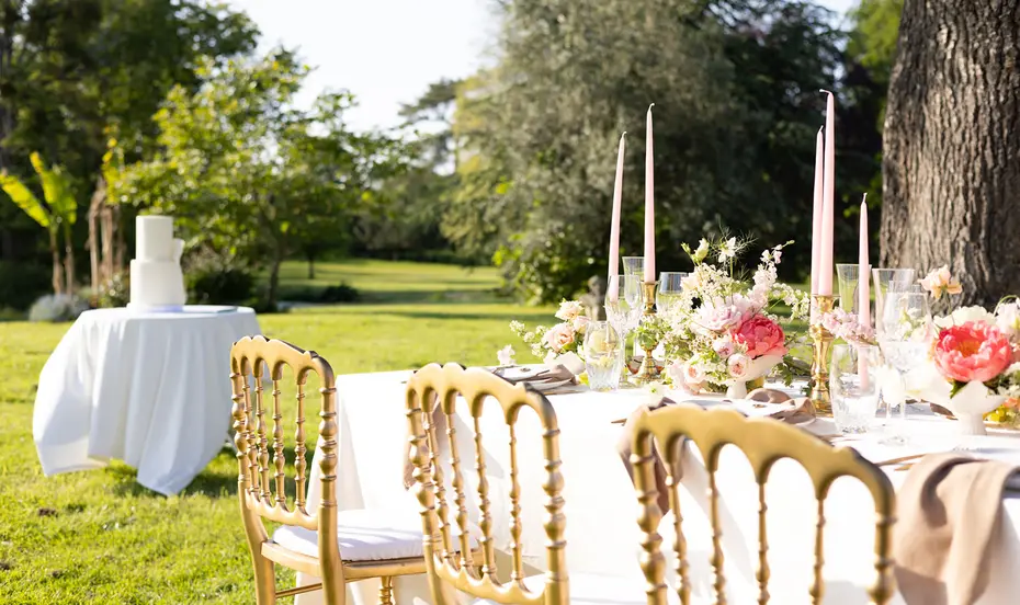 A table set beneath the trees, with gilded chairs, tall candles and a white cake