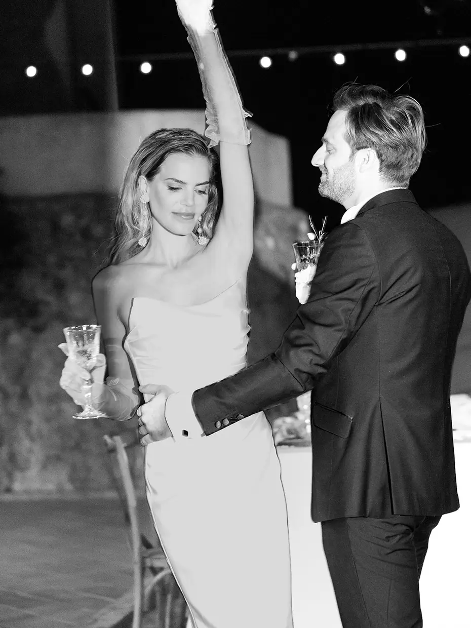 In black and white, a bride raising her glass with her arm outstretched, facing the groom under strings of lights