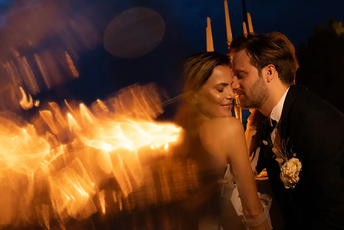 Bride and groom after dark, almost touching, the evening's firelight streaking past in the foreground.