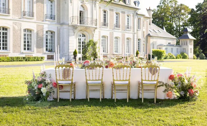 A long banquet table set on the lawn in front of a château façade, with floral runners and gilded chairs.