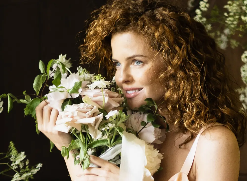 A guest in a pale pink dress breathing in a bouquet of white flowers, seen from the side against a dark background.