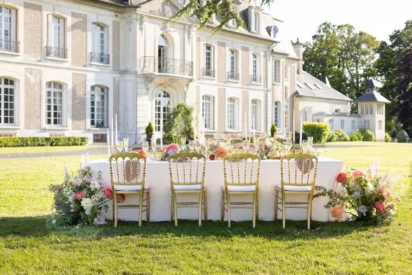 A long banquet table set on the lawn in front of a château façade, with floral runners and gilded chairs.