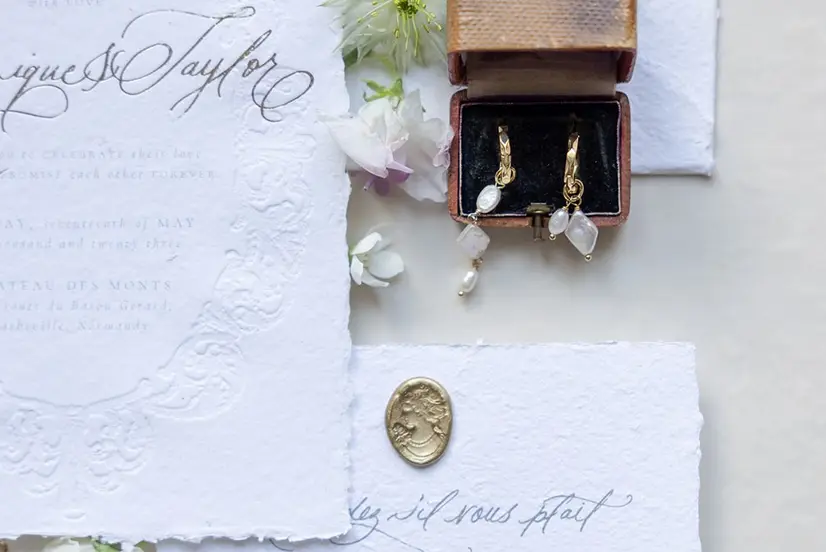 Calligraphed wedding stationery, a jewellery box holding earrings and a gold wax seal on textured paper.