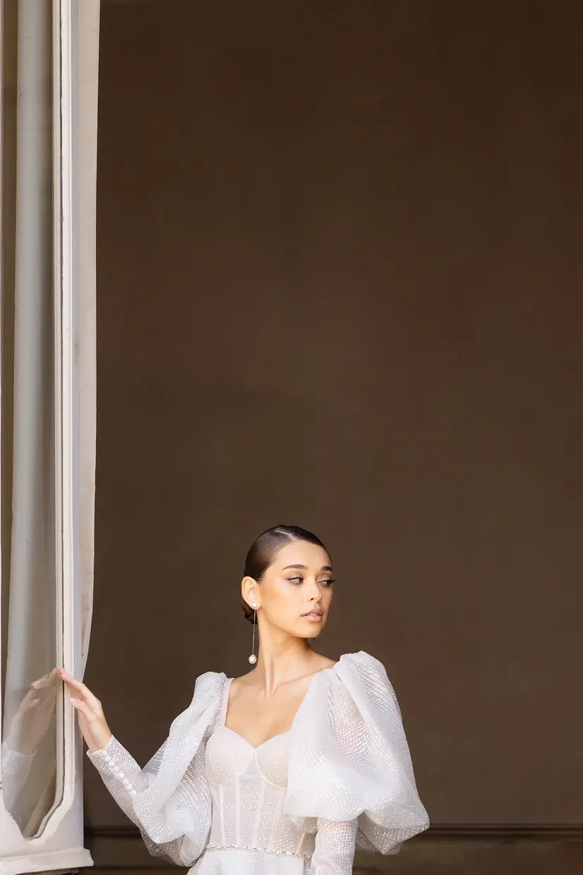 Studio portrait of a bride in a puff-sleeved gown, drawing back a pale curtain against a warm brown backdrop.