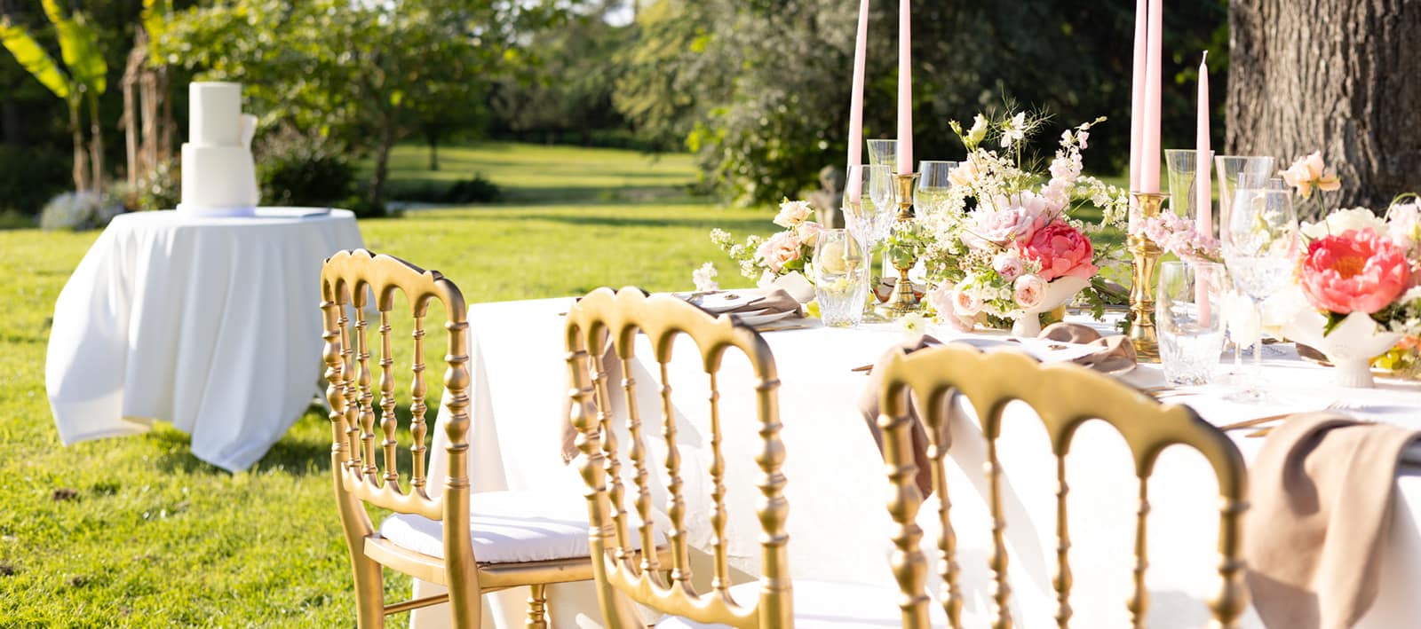 A garden reception table at golden hour: gilded chairs around white linen, tall pink tapers and peonies, the wedding cake on its own table on the lawn.